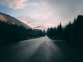 Beautiful shot of a black road surrounded by pine trees and mountains under a pinkish cloudy sky Royalty Free Stock Photo