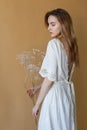 Beautiful skinny young girl with long hair in white dress on beige background holding white flowers in her hands