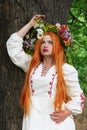 Beautiful slavic girl with long red hair with flower crown in white and red embroidered ethnic suit stands by tree Royalty Free Stock Photo