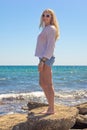 A beautiful slender blonde with long hair in a white shirt and shorts walks on the stones on the seashore Travel and