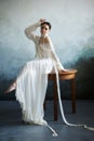 Beautiful slender brunette girl sitting on the sofa in a long white dress. Portrait of a woman with a jewellery on neck. Perfect Royalty Free Stock Photo