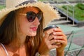 Beautiful slender tanned woman in a straw hat and a white sundress sits at a table on a summer terrace of a cafe and drinks coffee Royalty Free Stock Photo