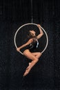 Beautiful female acrobat sitting in aerial hoop under rain on black background