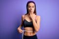 Beautiful slim girl controlling weight using measure tape on waist over purple background cover mouth with hand shocked with shame