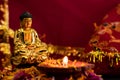 Beautiful small statue of buddha in maditation position with shining diya on flower petals against red golden background. religion Royalty Free Stock Photo