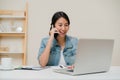 Beautiful smart business Asian woman in smart casual wear working on laptop and talking on phone while sitting on table in Royalty Free Stock Photo