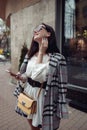 Beautiful smiled woman in stylish outlook standing at city street with smartphone in hand.