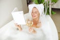 Beautiful smiled young woman lying in bath with foam, reading book and drinking wine.