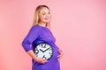 Beautiful and smiley blonde pregnant woman holding a clock in hands in studio on a pink background . time for birth Royalty Free Stock Photo