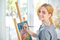 Young smiling girl paints on canvas with oil colors in own workshop. Window on the background. Art concept. Royalty Free Stock Photo