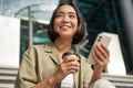 Beautiful smiling asian girl, drinking coffee, using mobile phone and sits on stairs outside. Young woman video chat Royalty Free Stock Photo