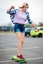 A beautiful smiling blond girl wearing checkered shirt, cap and denim shorts is balancing on the grin longboard and