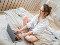 Beautiful smiling blonde girl sitting on bed and using laptop Royalty Free Stock Photo