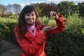 Beautiful smiling brunette girl in red coat holding fresh green apple on palm of her hand over nature background Royalty Free Stock Photo