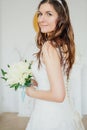 Beautiful smiling brunette woman bride in wedding dress with classical white roses bouquet in living room