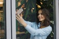 Beautiful smiling caucasian young woman is standing near the window and taking a selfie photo.