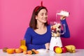 Beautiful smiling girl cooks smoothies in morning. Lots fruit on table. Charming woman adds milk to blender. Goodlooking female Royalty Free Stock Photo