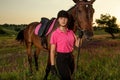 Beautiful smiling girl jockey stand next to her brown horse wearing special uniform on a sky and green field background Royalty Free Stock Photo