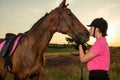 Beautiful smiling girl jockey stand next to her brown horse wearing special uniform on a sky and green field background Royalty Free Stock Photo