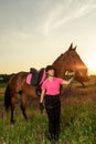 Beautiful smiling girl jockey stand next to her brown horse wearing special uniform on a sky and green field background Royalty Free Stock Photo