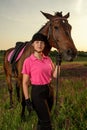 Beautiful smiling girl jockey stand next to her brown horse wearing special uniform on a sky and green field background Royalty Free Stock Photo