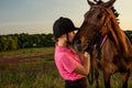 Beautiful smiling girl jockey stand next to her brown horse wearing special uniform on a sky and green field background Royalty Free Stock Photo