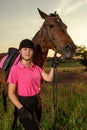 Beautiful smiling girl jockey stand next to her brown horse wearing special uniform on a sky and green field background Royalty Free Stock Photo