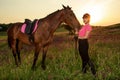 Beautiful smiling girl jockey stand next to her brown horse wearing special uniform on a sky and green field background Royalty Free Stock Photo