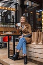 Beautiful smiling girl in trench coat and jeans joyfully looking in camera holding white cup of tea in hand while