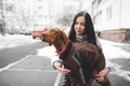 Beautiful smiling girl in warm winter clothes keeps a dressed dog on the background of the street and buildings. Walking with a Royalty Free Stock Photo