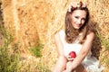 Beautiful smiling girl wearing white summer dress and floral head wreath sitting at the haystacks and holding a red apple Royalty Free Stock Photo