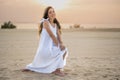 Young woman wearing white dress sitting on her kneed showing belly and looking over sunset on the beach. Royalty Free Stock Photo