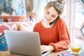 Beautiful smiling red-haired freckled girl working with laptop at the table restaurant during a conversation on the phone. Royalty Free Stock Photo