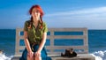 Beautiful smiling red-haired traveler girl sitting on a bench on the sea beach, dreams, relaxes and enjoys life. Royalty Free Stock Photo