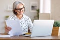 Smiling senior woman holding paper financial document and looking at laptop while working from home Royalty Free Stock Photo