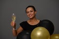 Beautiful smiling woman holding a glass with sparkling wine poses with golden and black inflated air balloons against gray wall Royalty Free Stock Photo