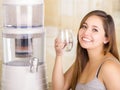 Beautiful smiling woman holding a glass of water, with a filter system of water purifier on a kitchen background