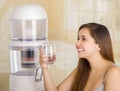 Beautiful smiling woman holding a glass of water, with a filter system of water purifier on a kitchen background