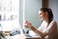 Beautiful smiling woman reading pleasant text message on mobile phone during work on laptop computer while sitting in modern coffe