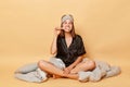 Beautiful smiling young girl sitting floor with blanket wearing black pajama and sleep eye mask isolated over beige background Royalty Free Stock Photo