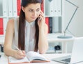 Beautiful smiling young girl talking on the mobile phone sitting at office and looking at laptop. Royalty Free Stock Photo