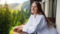 Beautiful smiling young woman in bathrobe looking on mountains and forest from the hotel room balcony Royalty Free Stock Photo