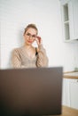 Beautiful smiling young woman fair long hair girl in glasses wearing in cozy knitted sweater using laptop at bright kitchen Royalty Free Stock Photo