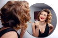 Beautiful smiling young woman with perfect make up, red lips, retro hairstyle in black dress, looking in mirror Royalty Free Stock Photo