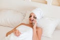 Beautiful smiling young woman with under eye patches and talking mobile phone in bathrobe lying in bed. Happy girl taking care of Royalty Free Stock Photo