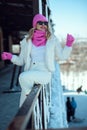 Beautiful smiling young woman wearing white puffer jacket and pants sitting on the wooden railing in the terrace of ski resort hot