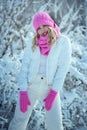 Smiling young woman wearing white puffer jacket and pants, pink knit hat, scarf and mittens standing in the snowy bushes