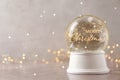 Beautiful snow globe with phrase Merry Christmas on marble table against festive lights, space for text