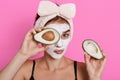 Beautiful spa woman with facial mask on face and holding halves of avocado in hands, looking at camera, doing cosmetology Royalty Free Stock Photo