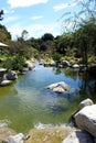 Beautiful stream lined with large rocks, plants and trees and a wooden bridge in the distance at the Japanese Friendship Garden